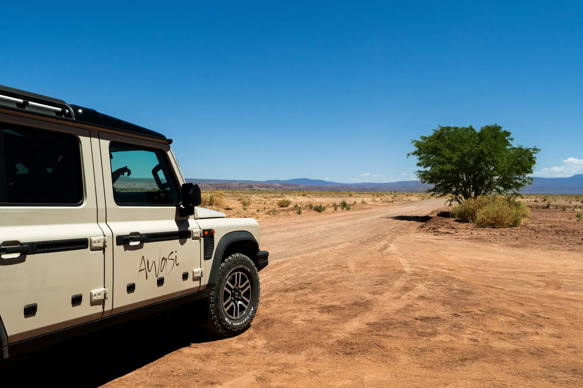Off-road suv parked beside a dusty desert road with a lone tree under a clear blue sky.