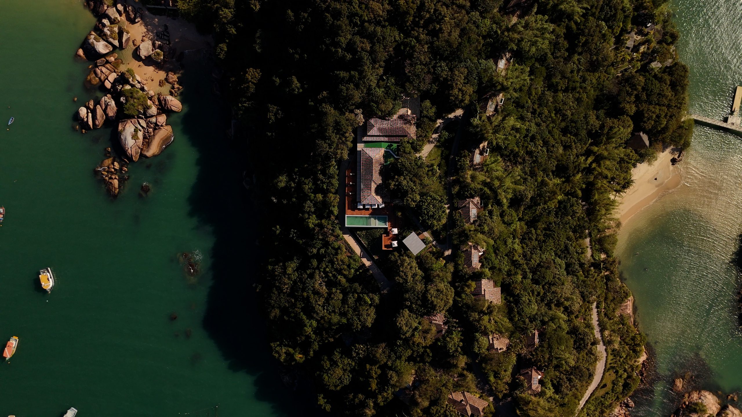 Aerial view of a luxury villa surrounded by lush forest and turquoise sea with rocky shoreline and small boats nearby