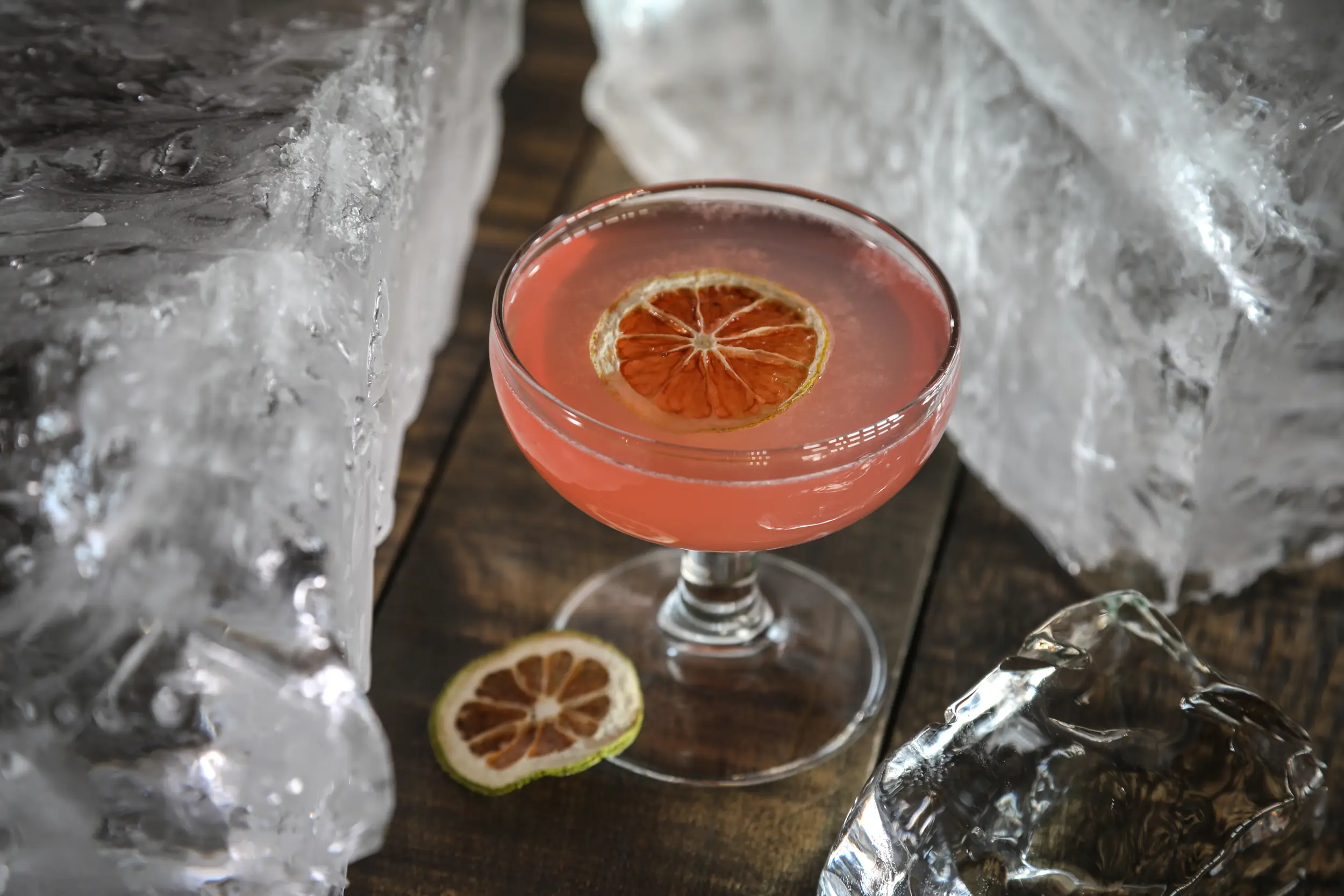 Pink Citrus Cocktail Garnished With A Dried Lime Slice, Surrounded By Large Blocks Of Ice On A Wooden Table.
