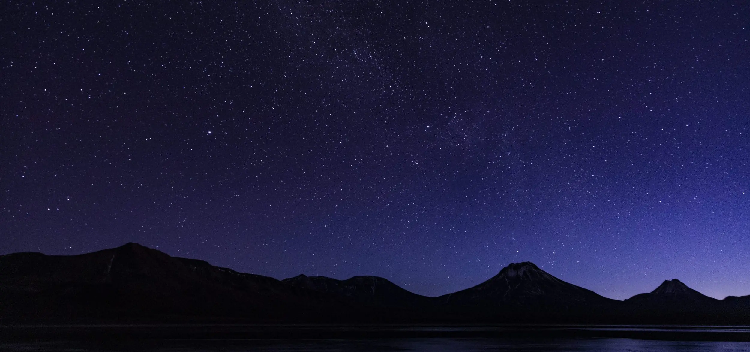 Star-filled night sky above dark mountain silhouettes reflected on a calm lake.