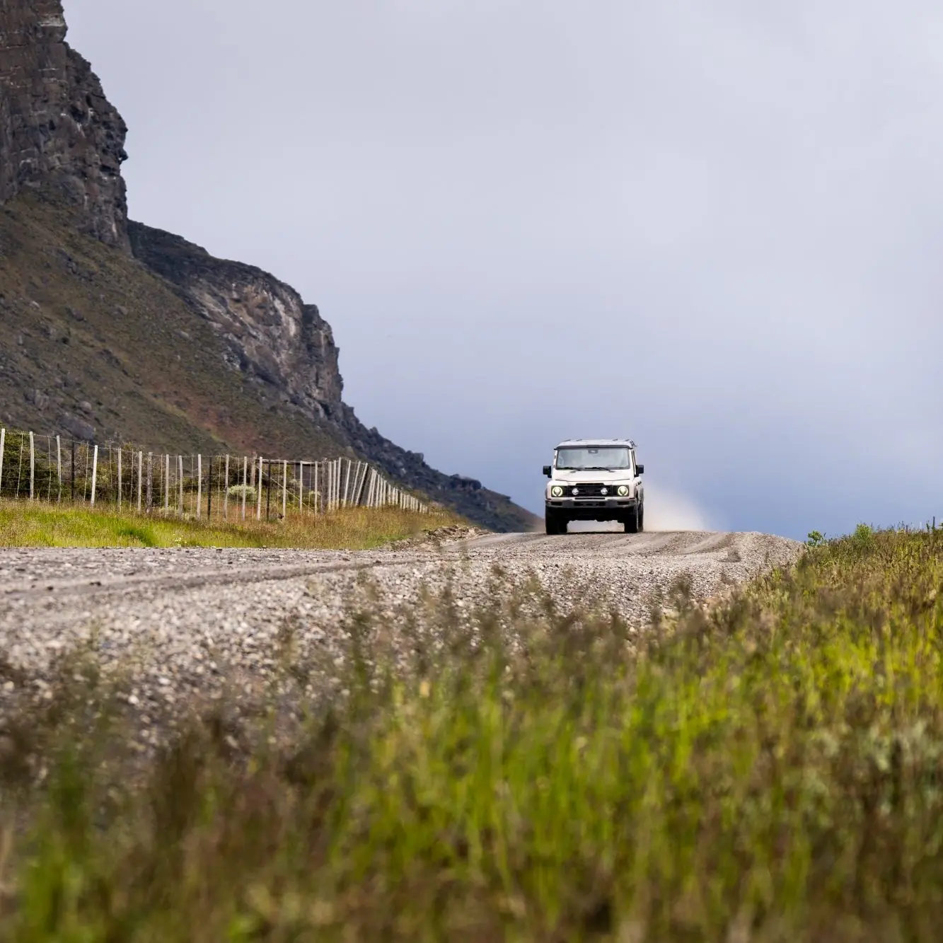 Suv driving along a gravel road beside rocky hills and grassy fields under an overcast sky.