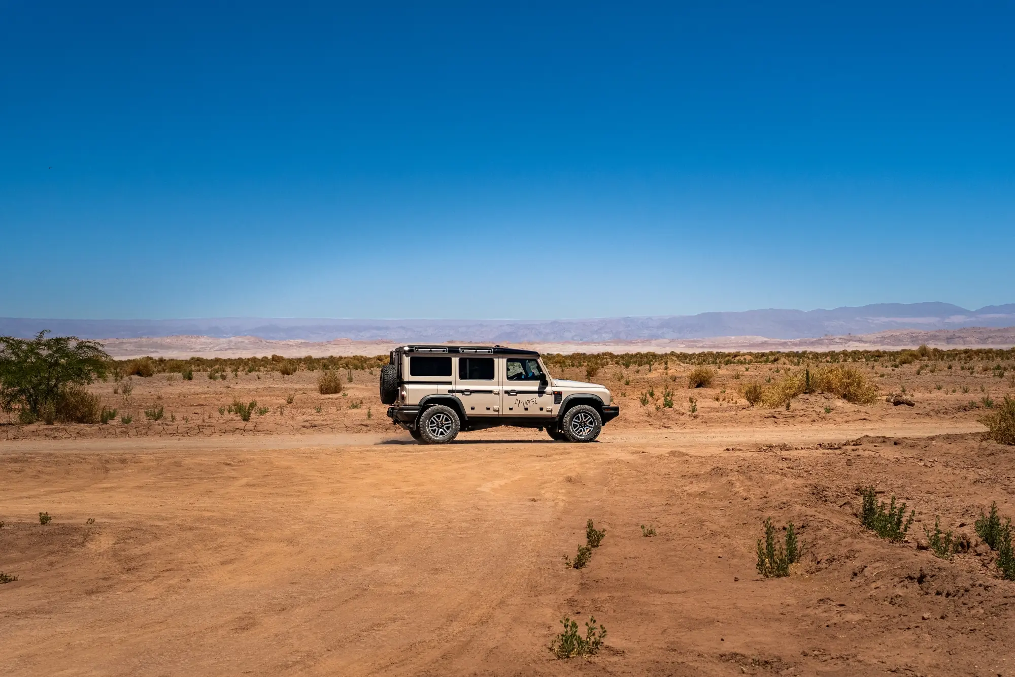 Off-road suv driving across a wide desert landscape beneath a clear blue sky.