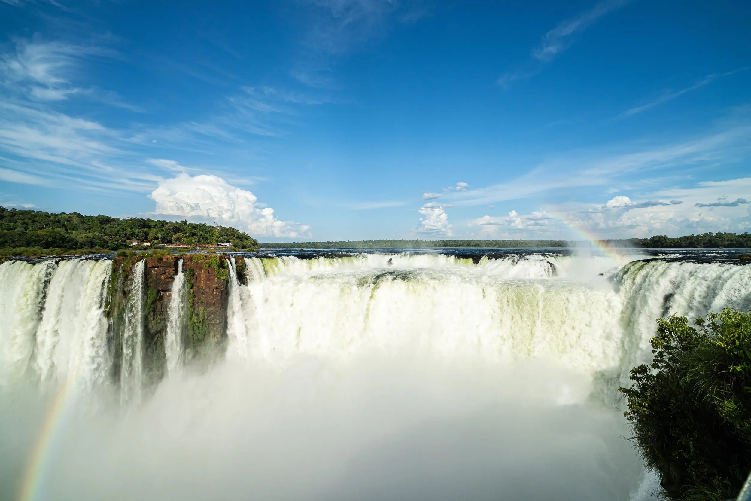 Massive Waterfall Cascading Into Mist With Rainbows Visible Beneath A Bright Blue Sky.
