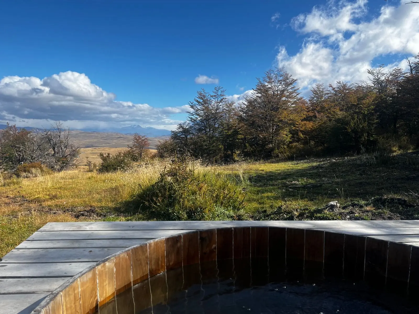 Wooden Outdoor Hot Tub Overlooking Grassy Hills, Trees, And Distant Mountains Beneath A Bright Blue Sky.
