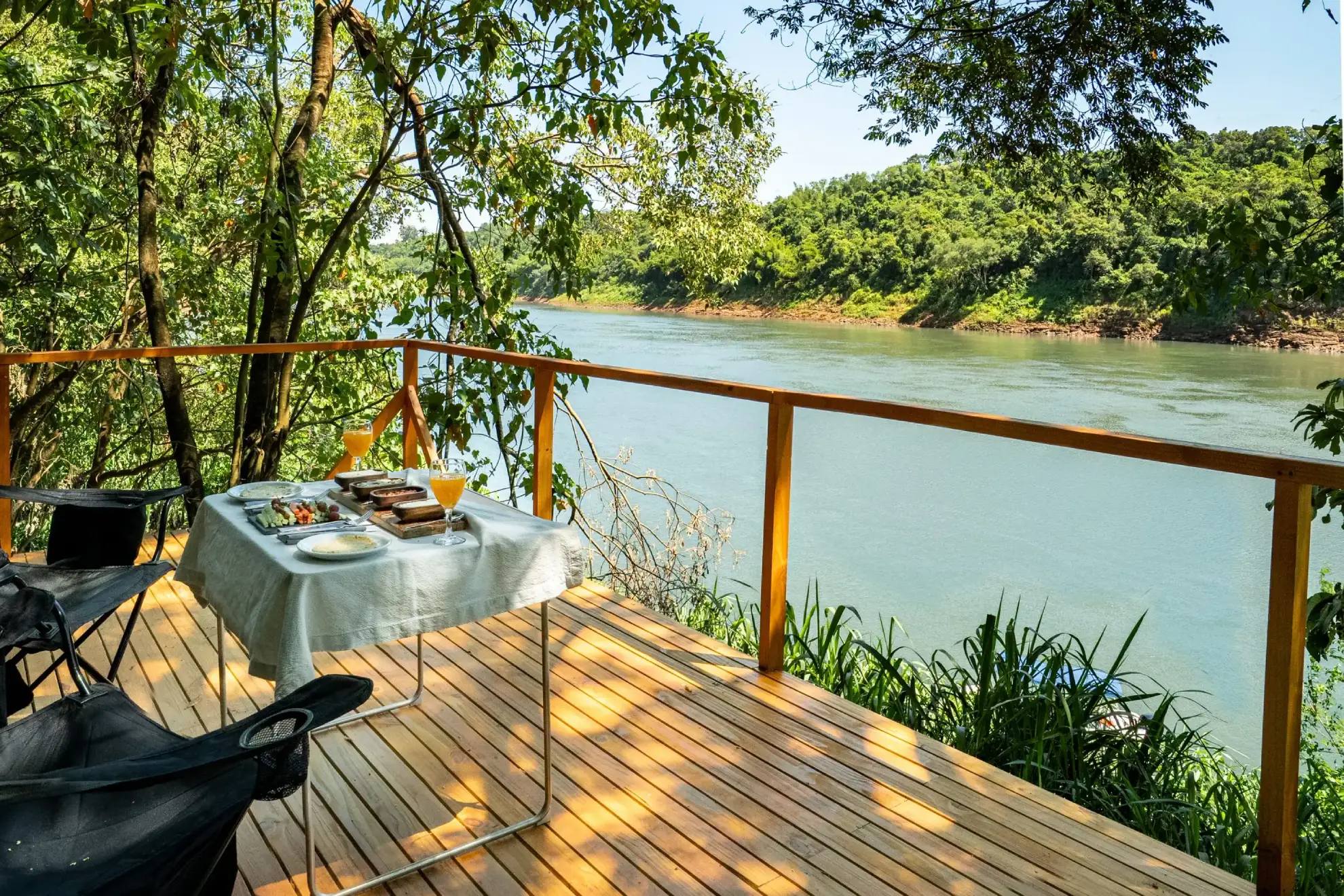 Riverside Deck Dining Setup Surrounded By Lush Forest With Drinks And Plates Overlooking Calm Water.
