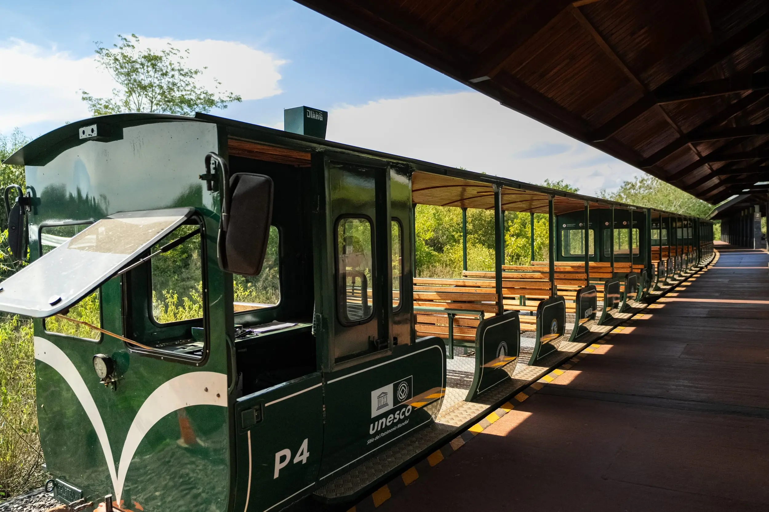 Open-Air Green Train At A Station Platform Surrounded By Lush Vegetation And Sunlight.