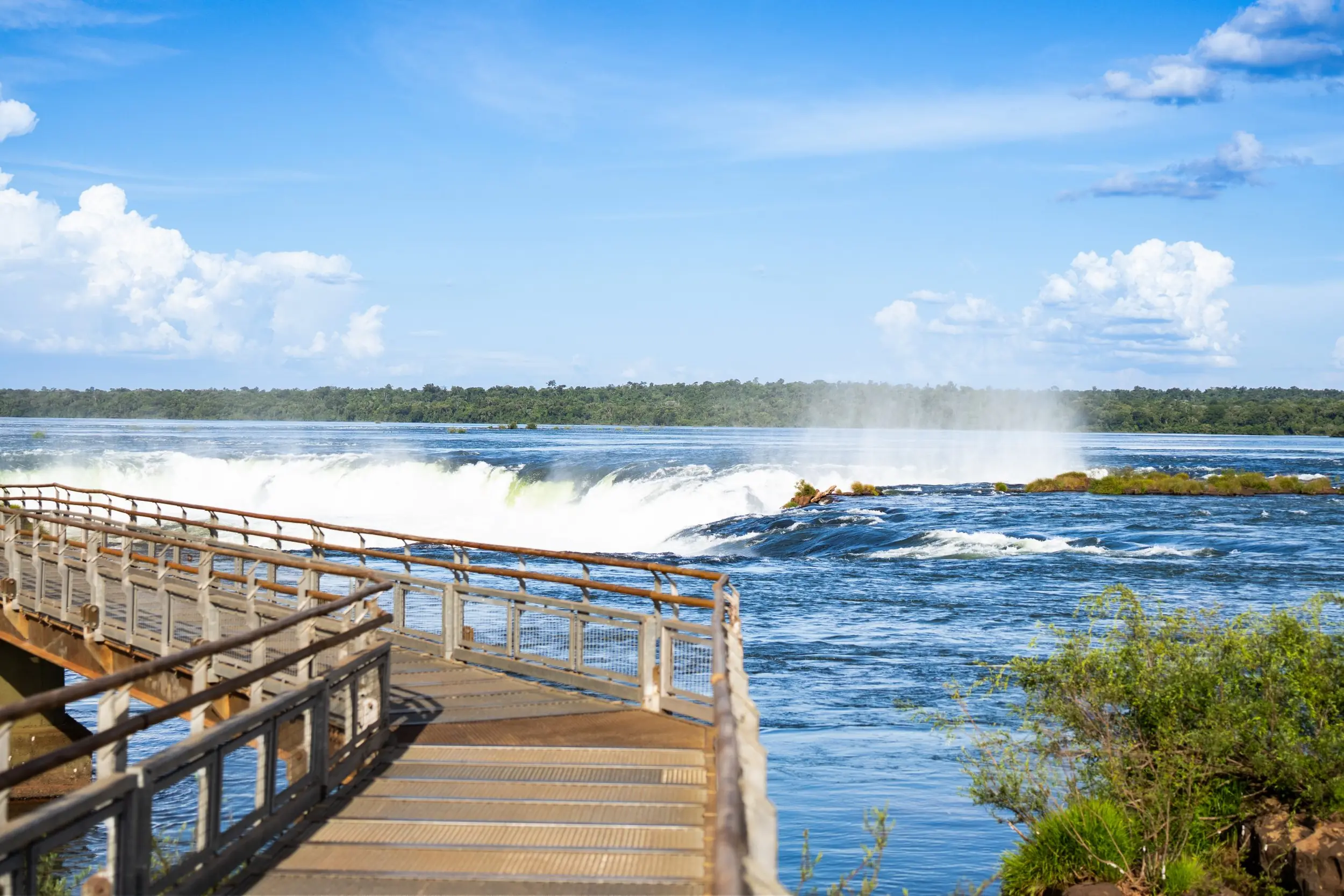 Wooden Boardwalk Overlooking A Wide Waterfall And Rushing River Under A Bright Blue Sky.