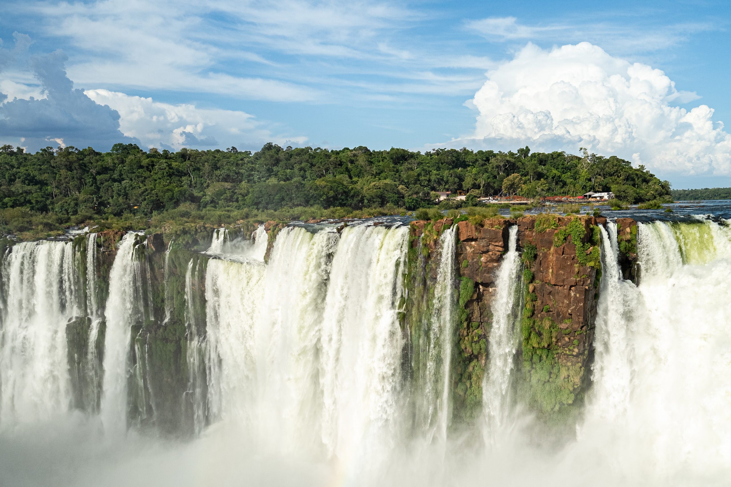Massive waterfall cascading over lush cliffs into mist beneath a bright blue sky with towering clouds.