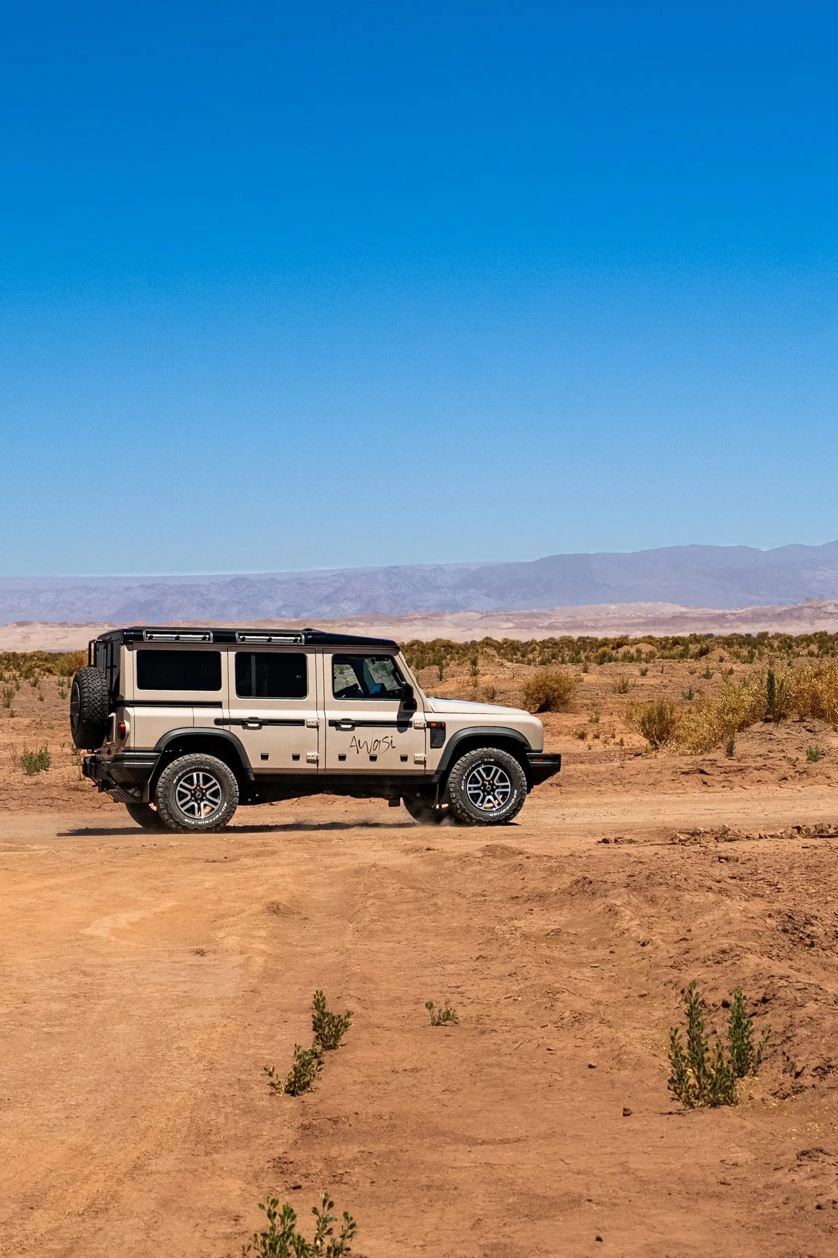 White off-road suv driving across a dusty desert landscape beneath a clear blue sky.