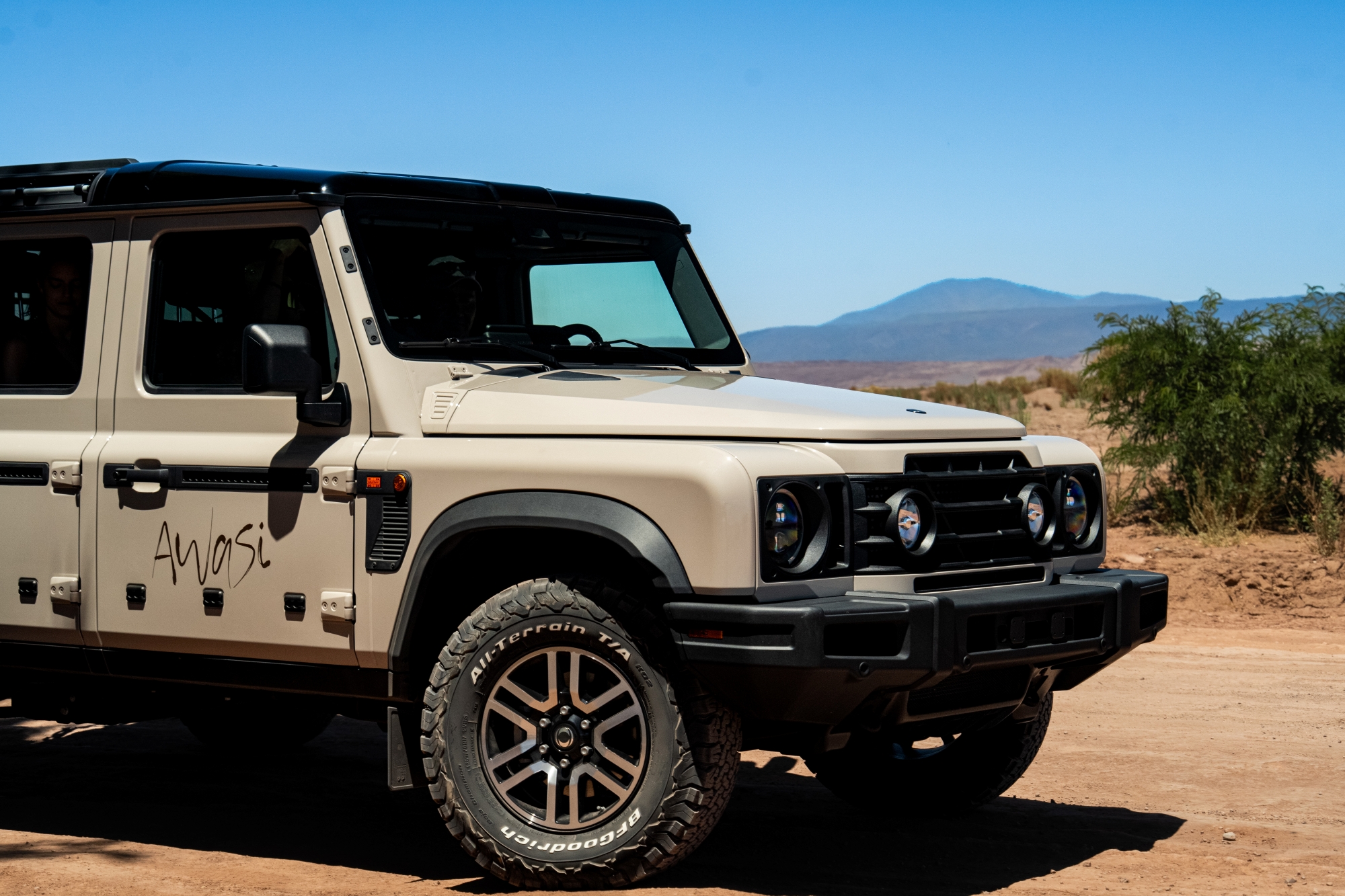 Beige Off-Road Suv Parked In A Desert Landscape With Mountains And Clear Blue Skies In The Background.