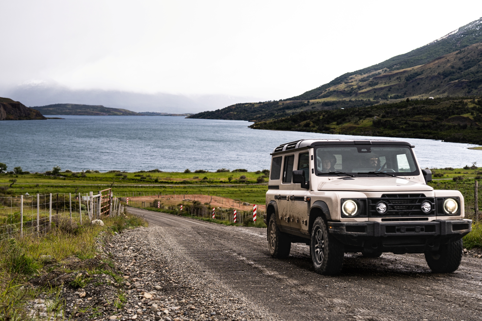 Off-Road Suv Parked On A Gravel Road Beside A Lake And Rolling Green Hills Under Cloudy Skies.