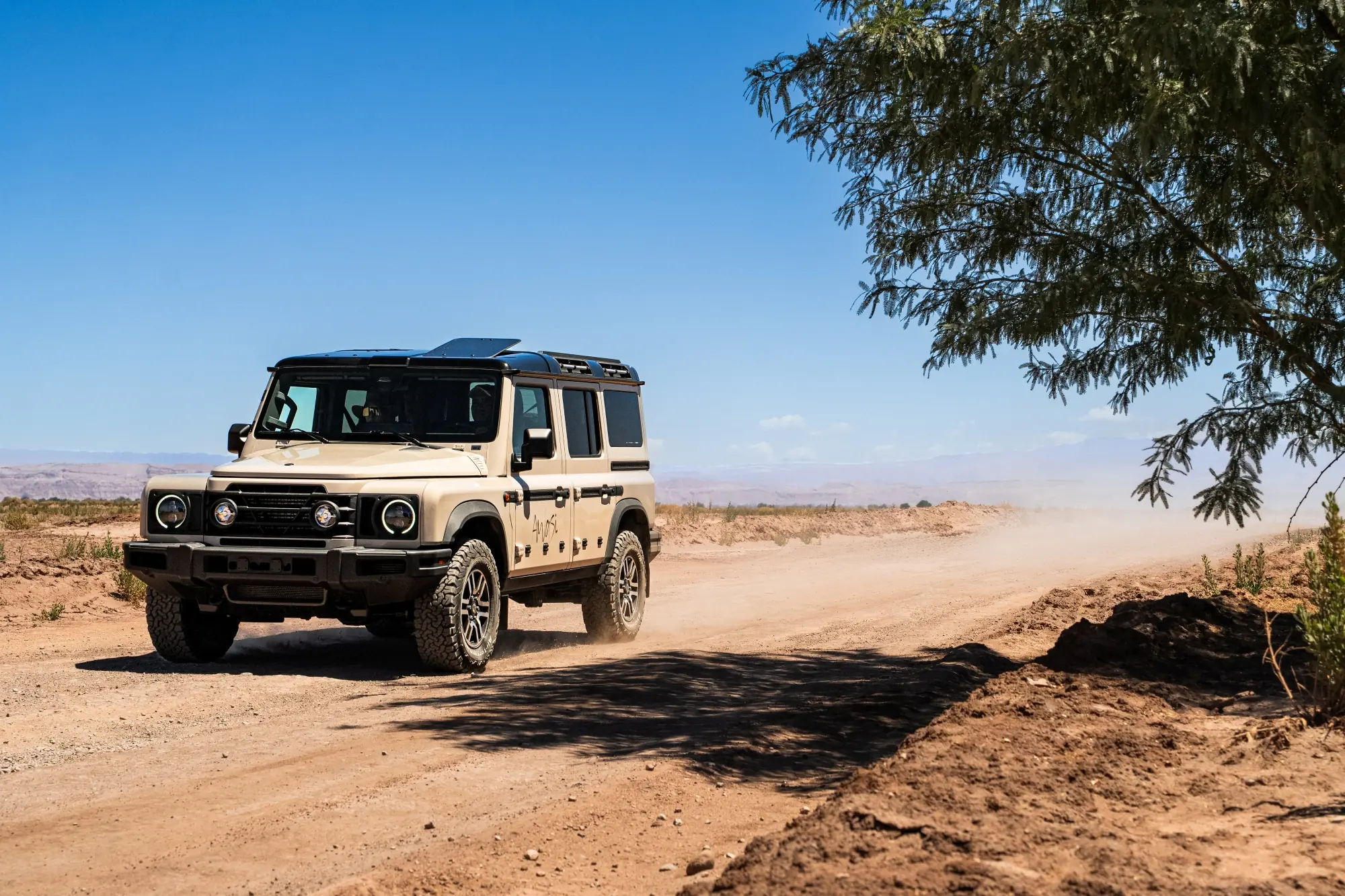 Beige off-road suv driving along a dusty desert road beside a tree under a clear blue sky.