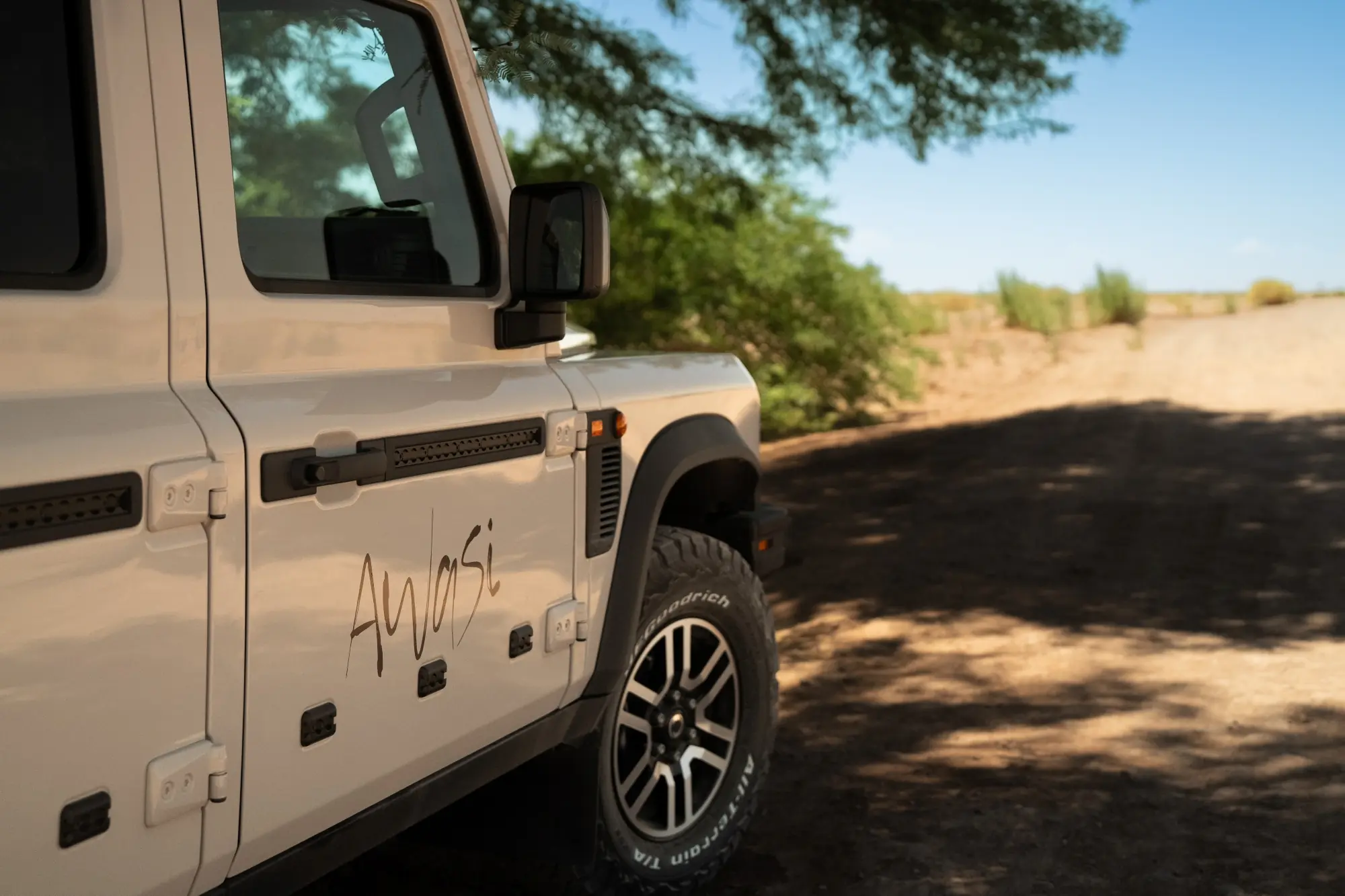 White off-road suv parked beside a sunlit dirt road lined with trees and desert landscape.