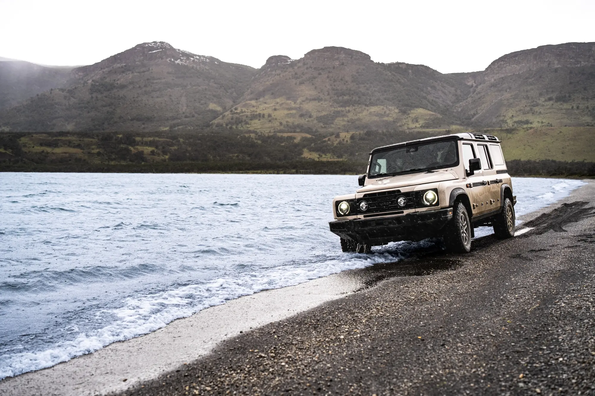 Off-road suv driving along a lakeshore with rugged mountains rising in the background.