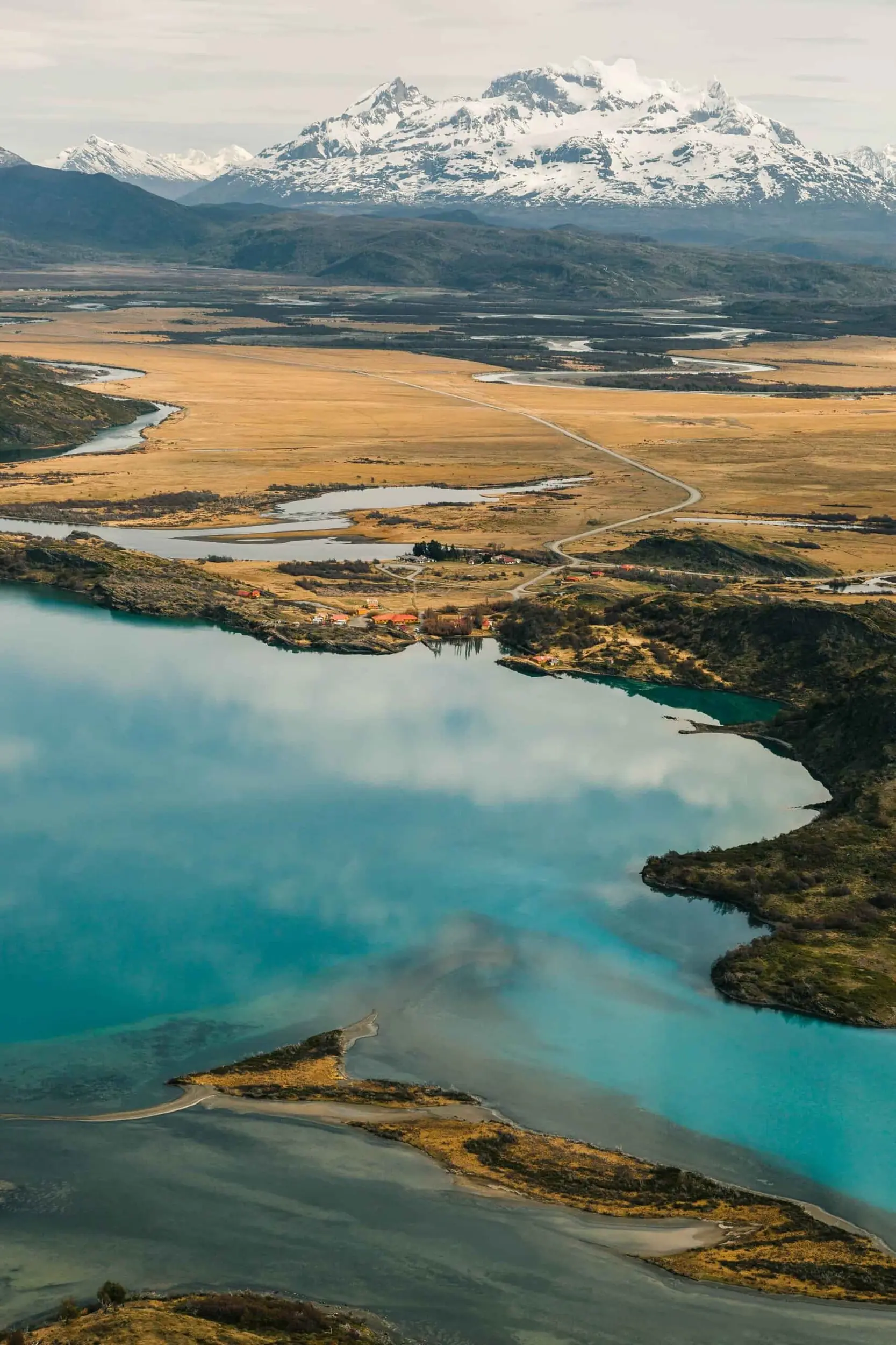 An Aerial View Of A Turquoise Lake And Vast Plains Before Snowy Mountains.