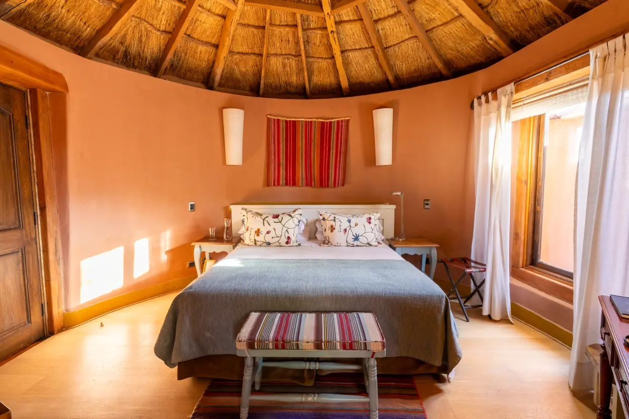 A Rustic, Circular Bedroom Features A Bed With Floral Pillows Beneath A Thatched Ceiling.