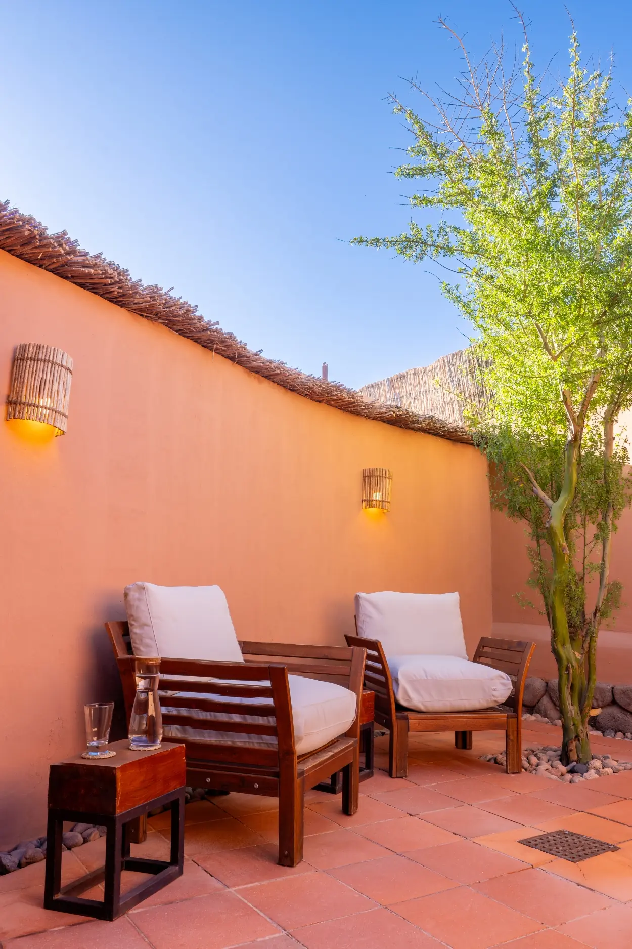 Two Wooden Lounge Chairs With White Cushions Sit On A Tiled Patio Next To A High Adobe Wall With Warm Sconce Lighting.