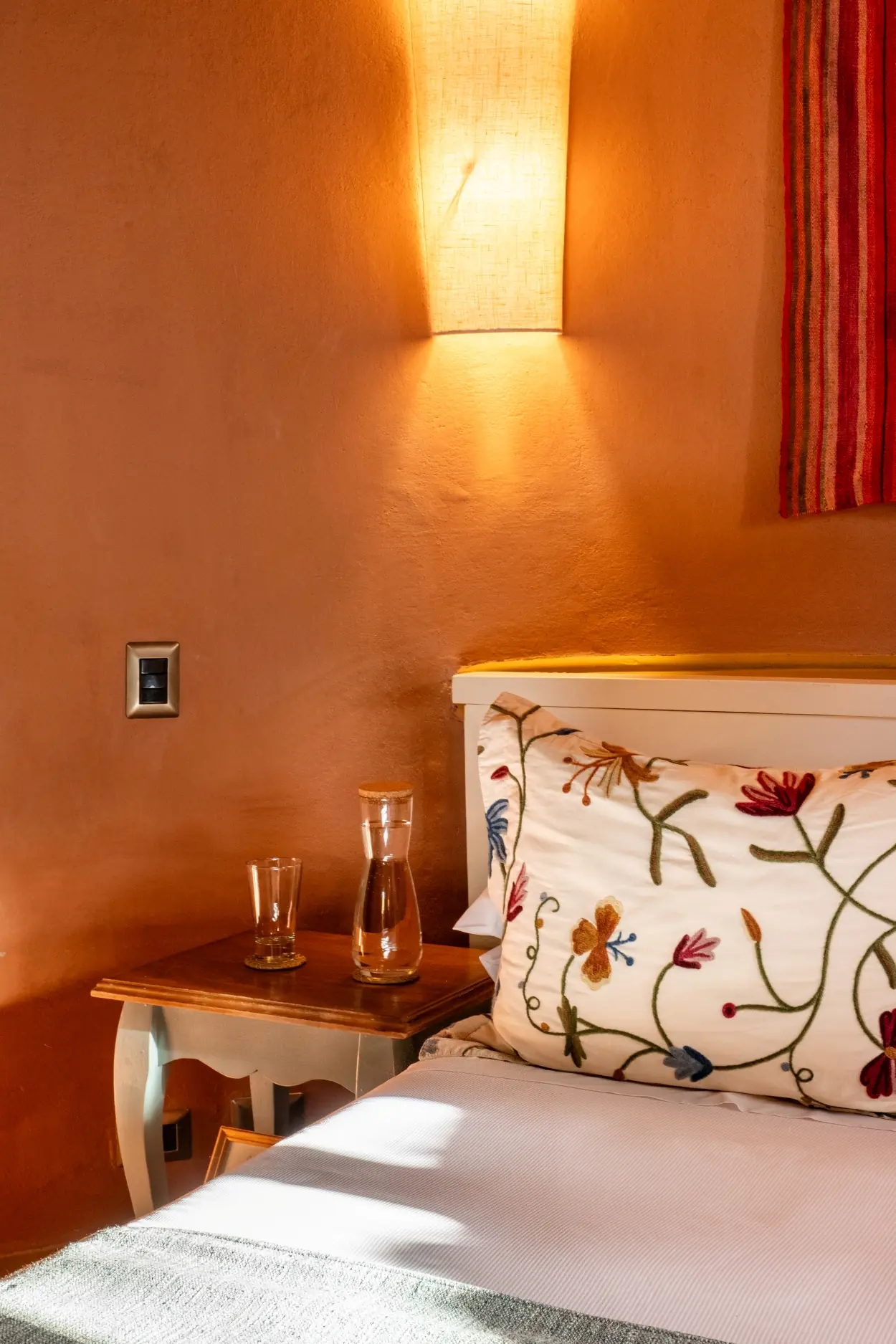 A Detailed Floral-Embroidered Pillow Rests On A Bed Next To A Wooden Nightstand With A Water Carafe And Glass Under A Warm Wall Sconce.