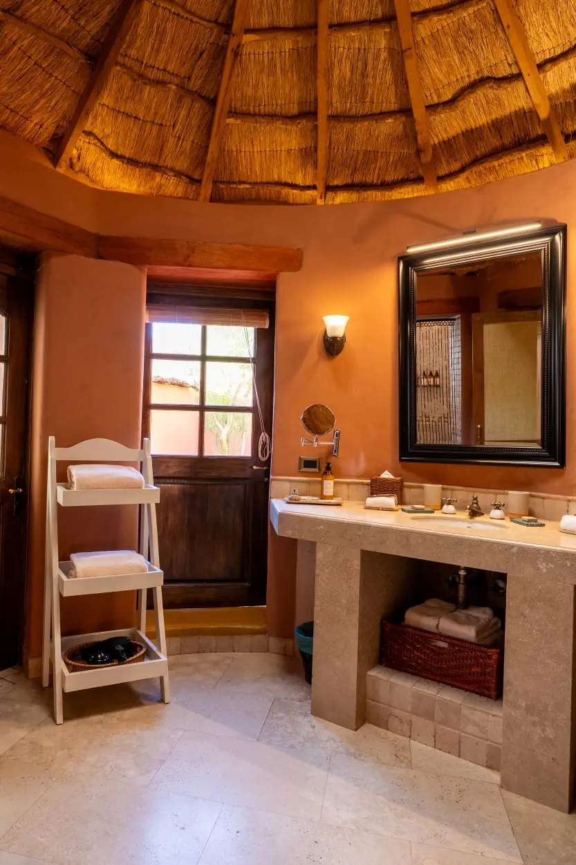 A Rustic, Stone-Countertop Vanity Stands In A Warm-Toned Bathroom Beneath A Thatched Ceiling And Large Framed Mirror.