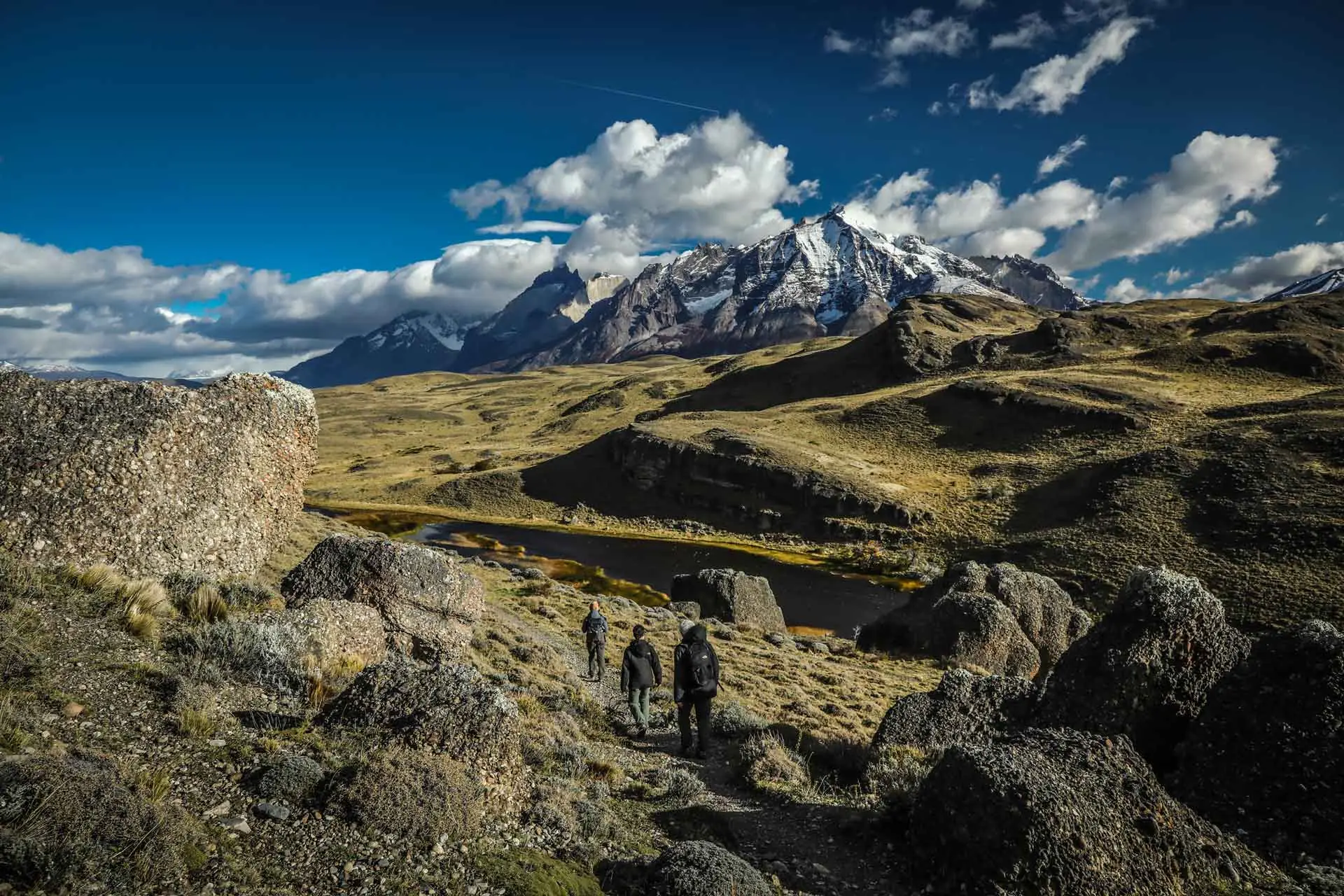 Three Hikers Walk Along A Rocky Path Toward Snow-Capped Mountains Under Puffy Clouds.