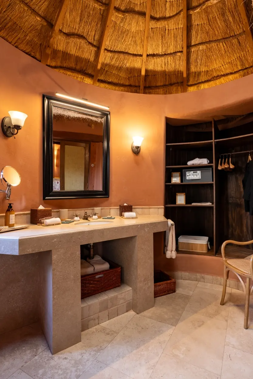 A Stone Vanity With A Large Black-Framed Mirror Sits Beneath A Thatched Ceiling, Adjacent To An Open Wooden Closet Alcove.