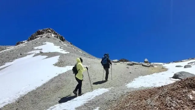 Two Hikers Using Trekking Poles Climb A Snowy Mountain Slope Beneath A Clear Blue Sky.
