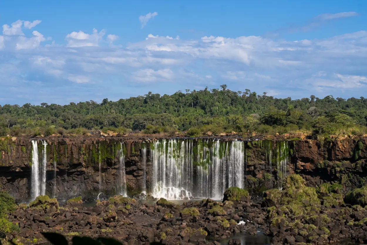 Wide waterfall cascading over rocky cliffs beneath a lush green forest and bright blue sky.
