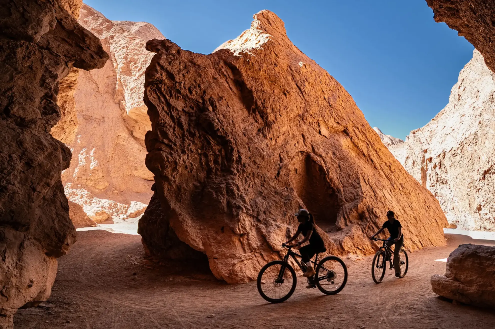 Two People Ride Mountain Bikes Through A Narrow, Sunlit Desert Rock Canyon.