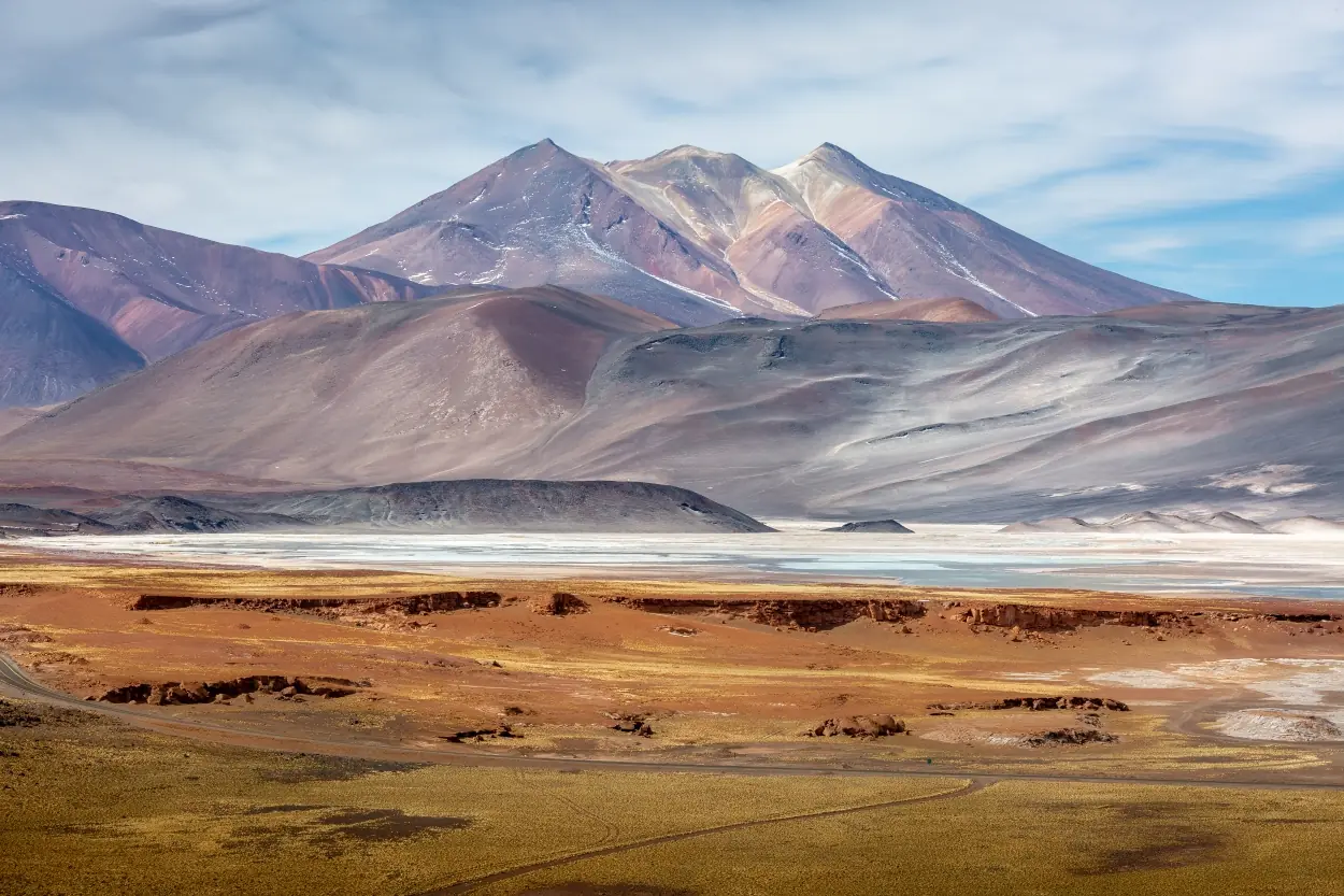 Pastel-Colored Mountain Range And Desert Landscape With A Shallow Lake Under A Cloudy Sky.