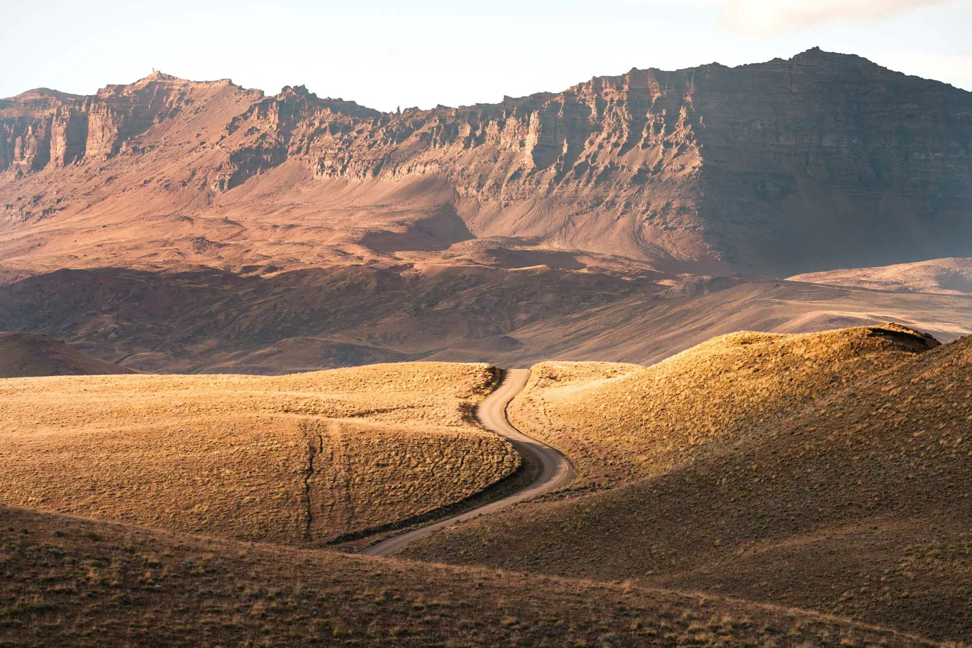 A Winding Dirt Road Cuts Through Rolling Golden Hills Toward Rugged, Sunlit Mountains.