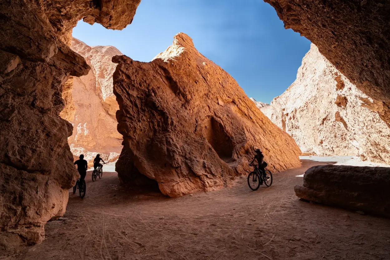 Cyclists riding through a dramatic desert canyon with towering red rock formations under a clear blue sky.