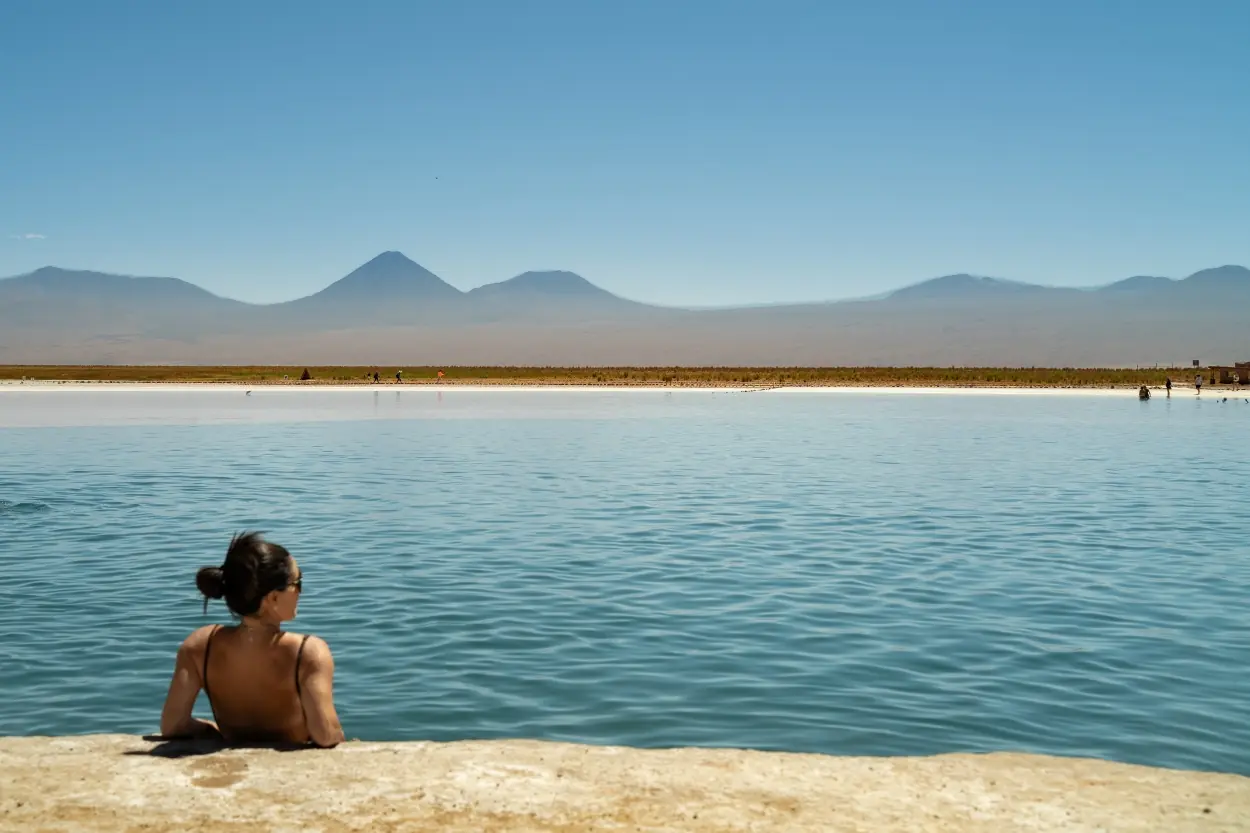 Person Relaxing At The Edge Of A Tranquil Lagoon With Mountains And Clear Blue Sky In The Distance.