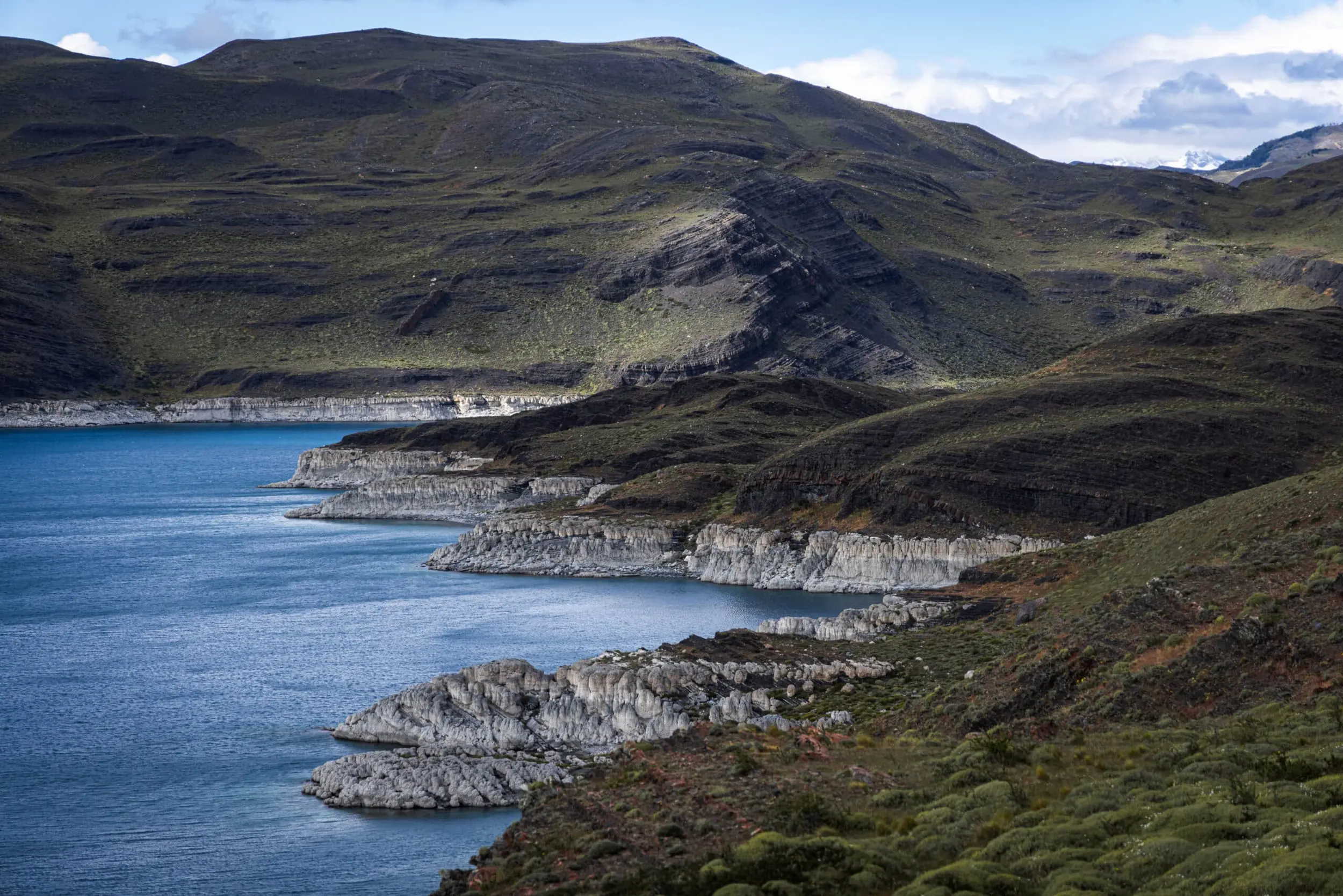 A Vibrant Blue Lake Curves Along Rugged, Rocky Hillsides Under A Soft Cloudy Sky.