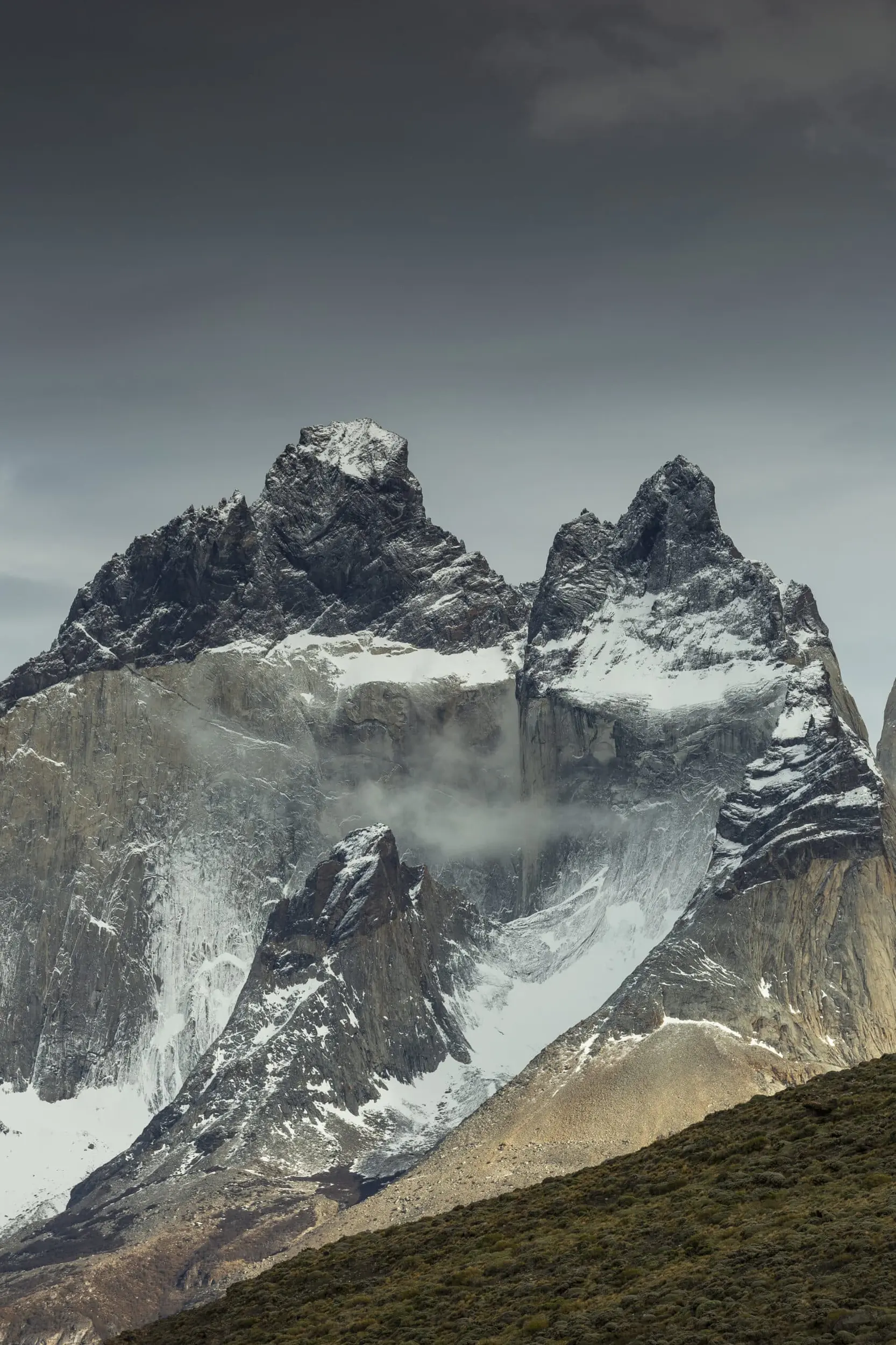 A Massive, Snow-Dusted Jagged Mountain Peak Stands Tall Under A Dramatic Overcast Sky.