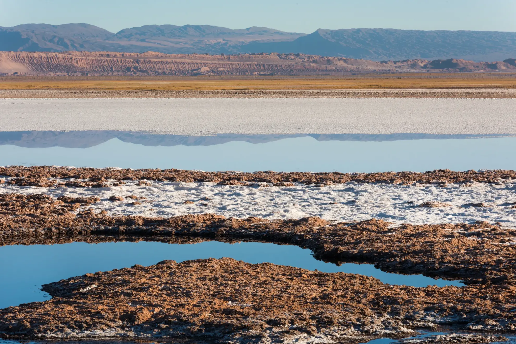 A Vast Salt Flat Features Calm Blue Water Reflecting Distant Mountains And Desert Landscapes.