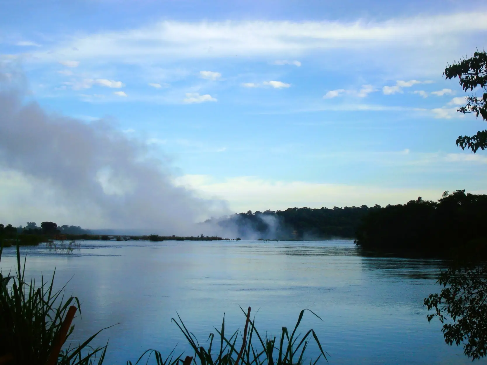 Mist Rising Over A Calm River Bordered By Dense Forest Beneath A Bright Blue Sky With Scattered Clouds.