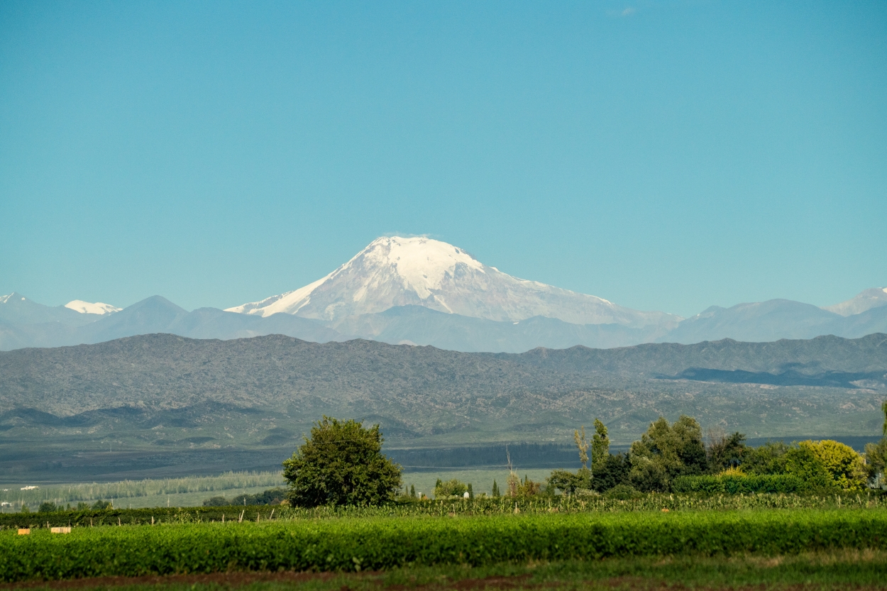 Snow-Capped Mountain Rising Above Green Fields And Rolling Hills Under A Clear Blue Sky.