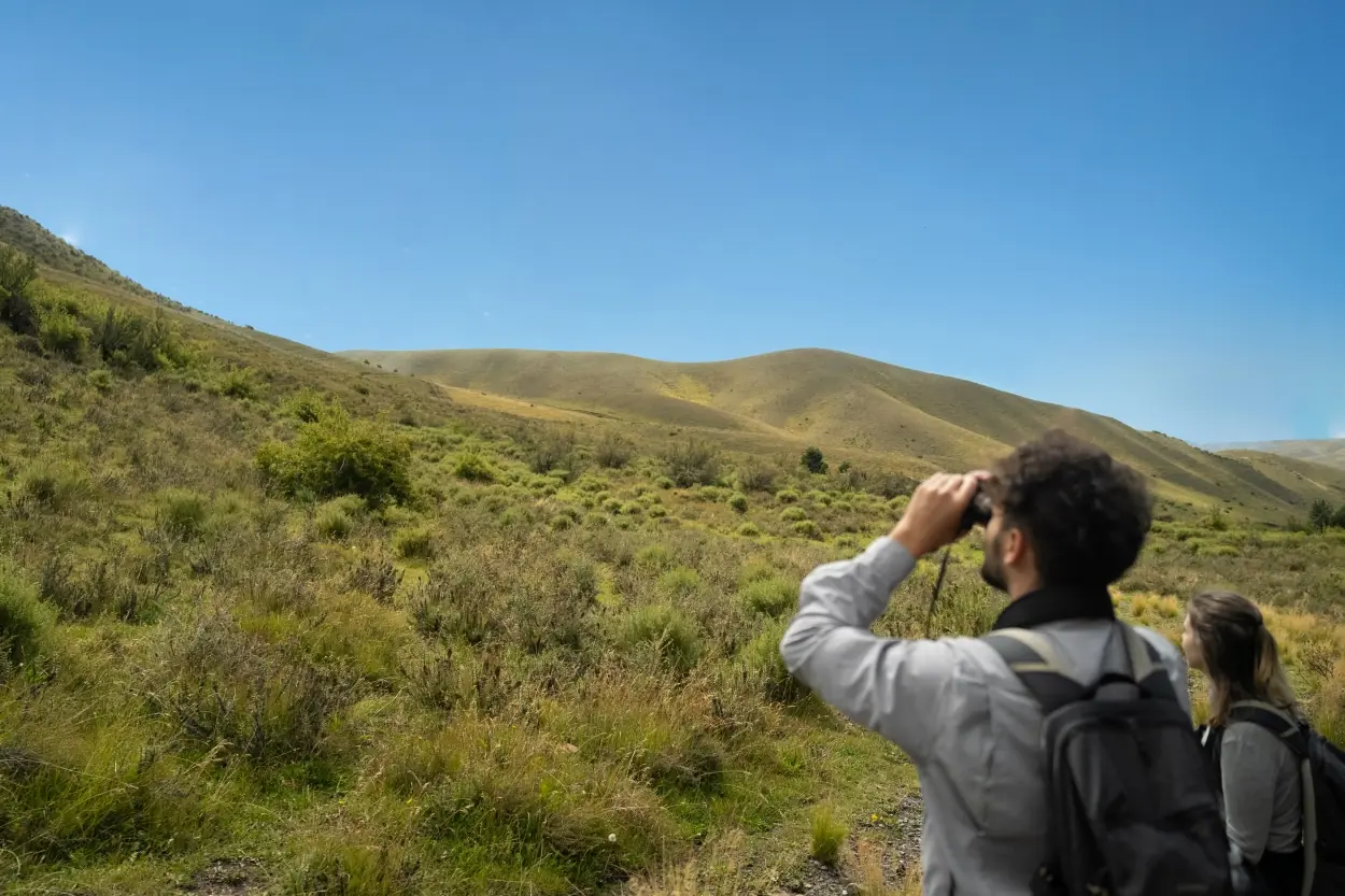 Two Hikers With Backpacks Using Binoculars In A Grassy Hillside Landscape Under A Clear Blue Sky.