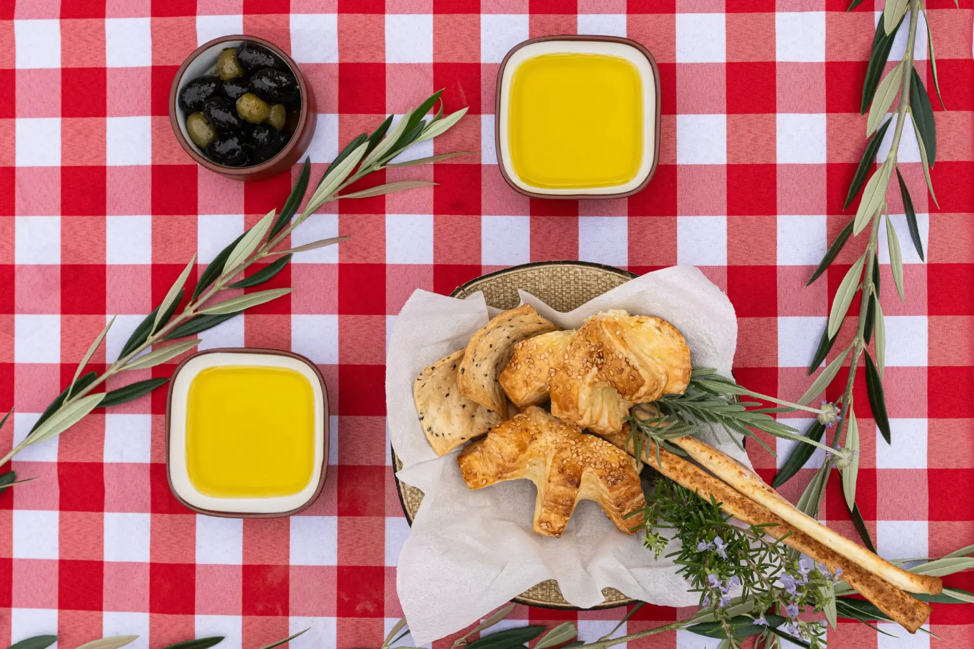 An Overhead View Of A Basket Of Pastries, Breadsticks, Olive Oil, And Olives Arranged On A Red-And-White Checkered Tablecloth.