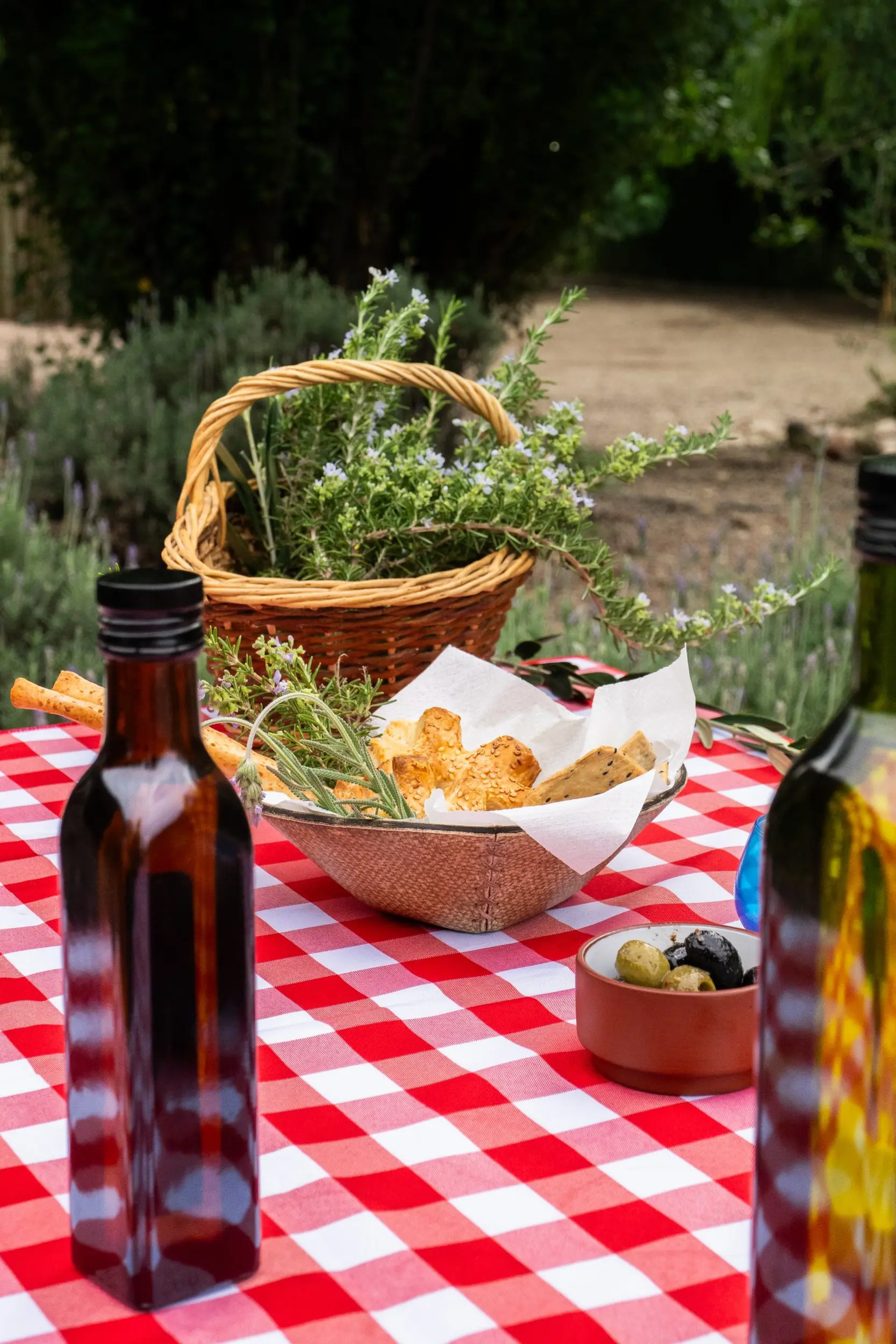 A Small Bowl Of Olives, A Basket Of Baked Goods, And Glass Bottles Sit On A Red-And-White Checkered Tablecloth Next To A Basket Of Flowering Rosemary.