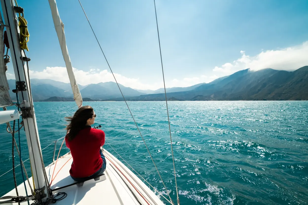 Person Sitting On A Sailboat Deck Overlooking Bright Blue Water And Distant Mountains On A Sunny Day.