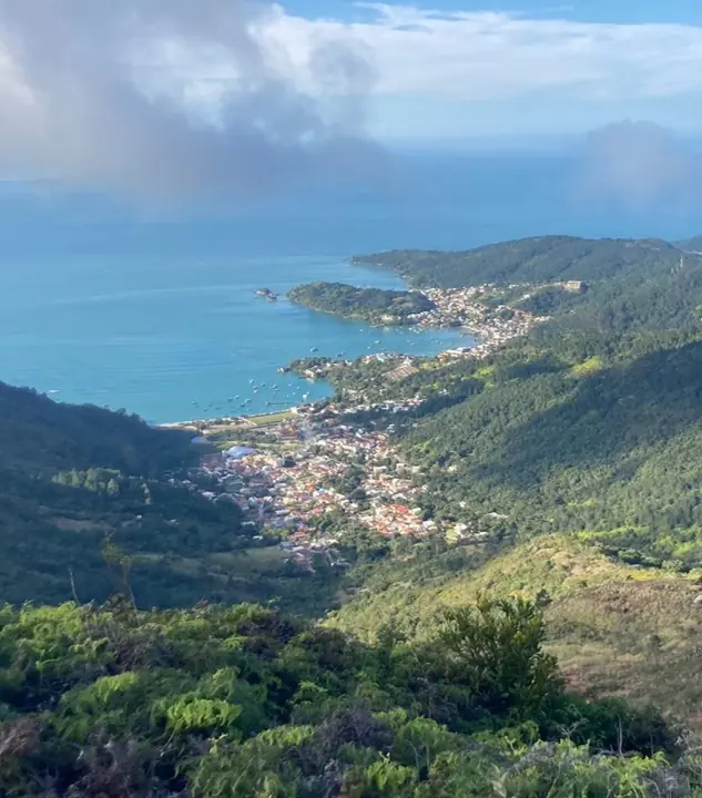 Aerial View Of A Coastal Town Between Lush Green Mountains And Blue Ocean Under Cloudy Skies.