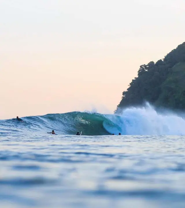 Surfers Waiting In The Ocean Near A Breaking Wave Beside A Lush Tropical Coastline At Sunset.