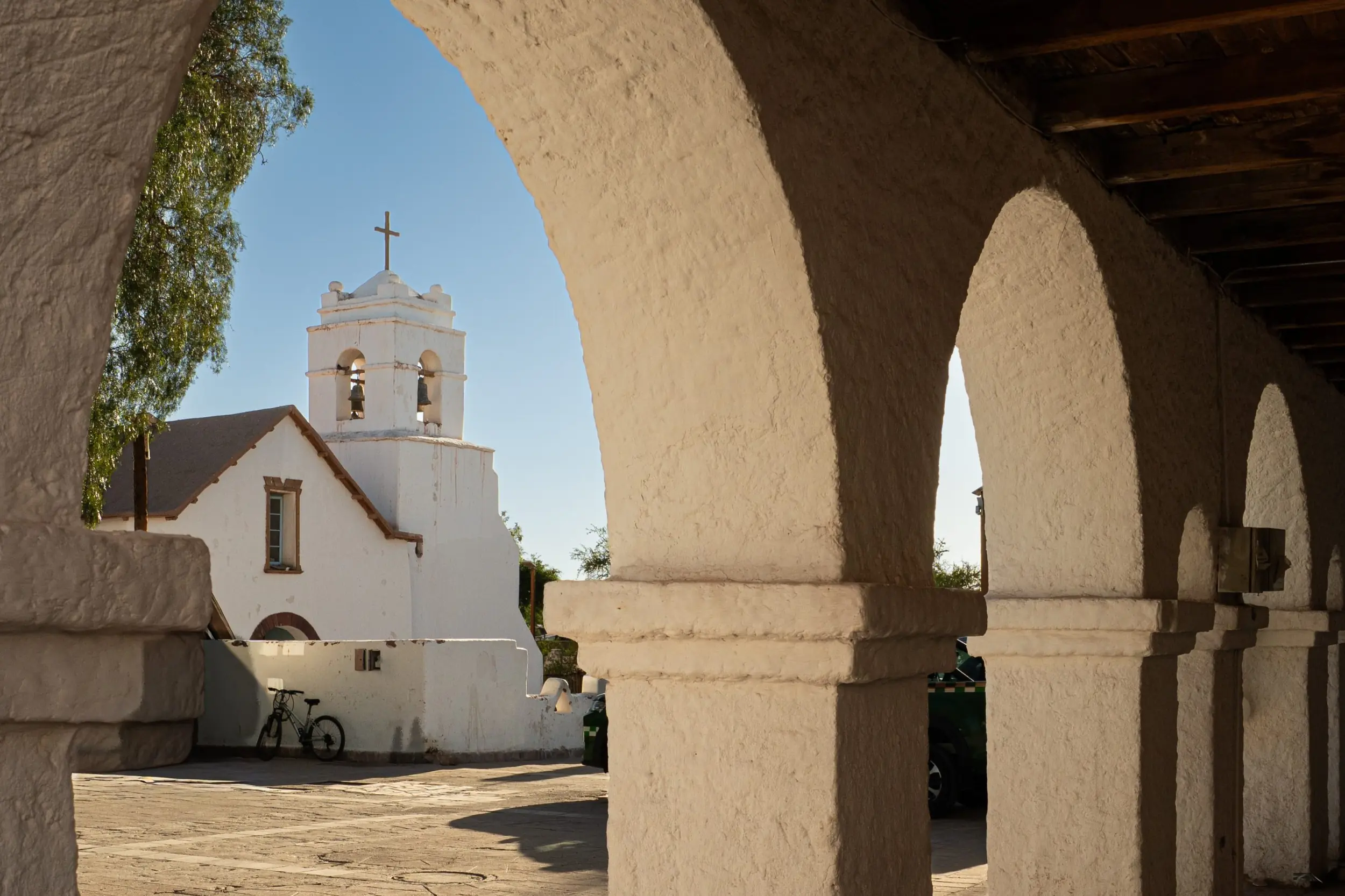 White Stucco Arches Frame A View Of A Historic Church Tower With A Cross.