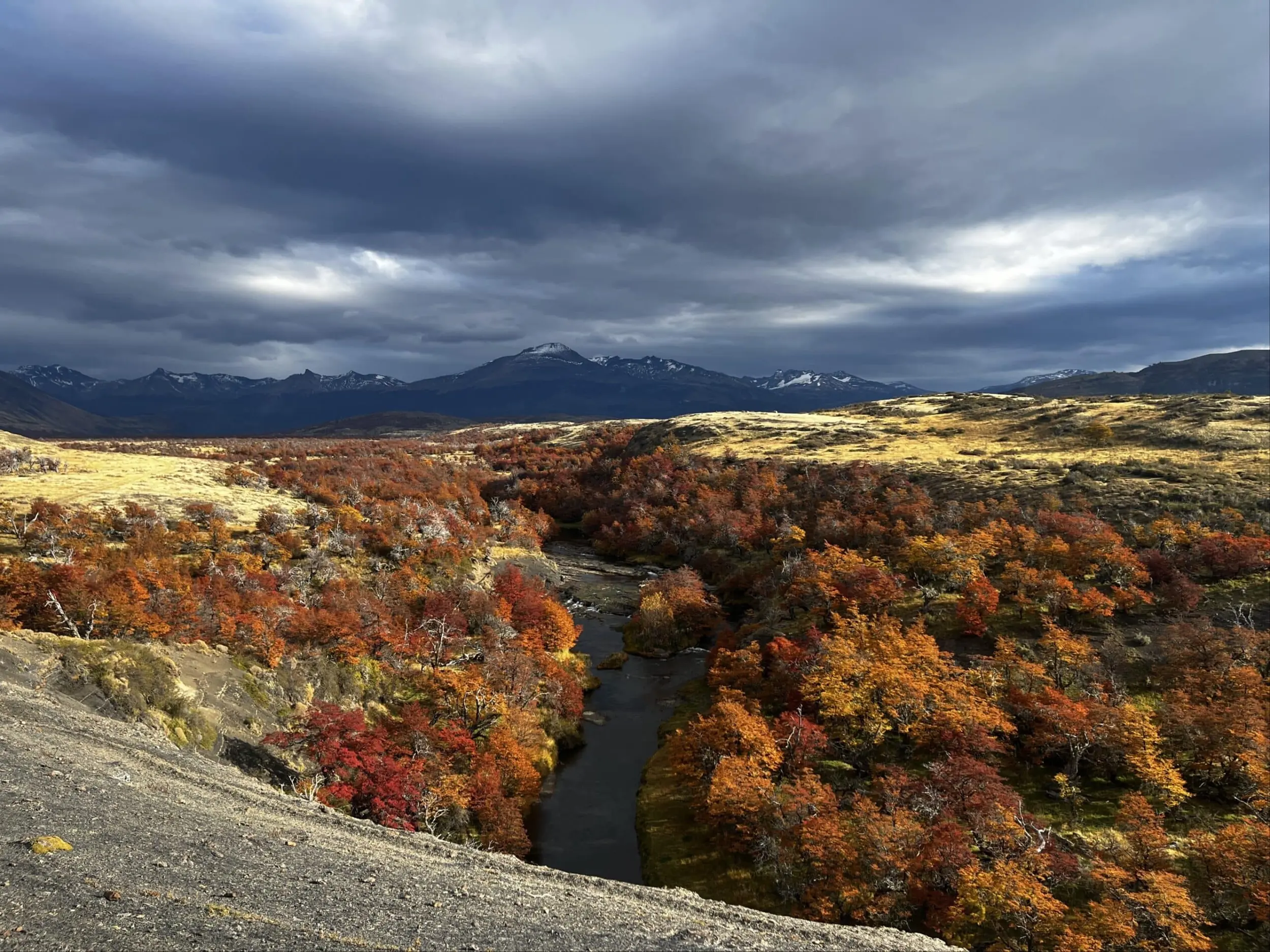 A River Winds Through Colorful Autumn Trees Below A Dramatic, Stormy Mountain Sky.