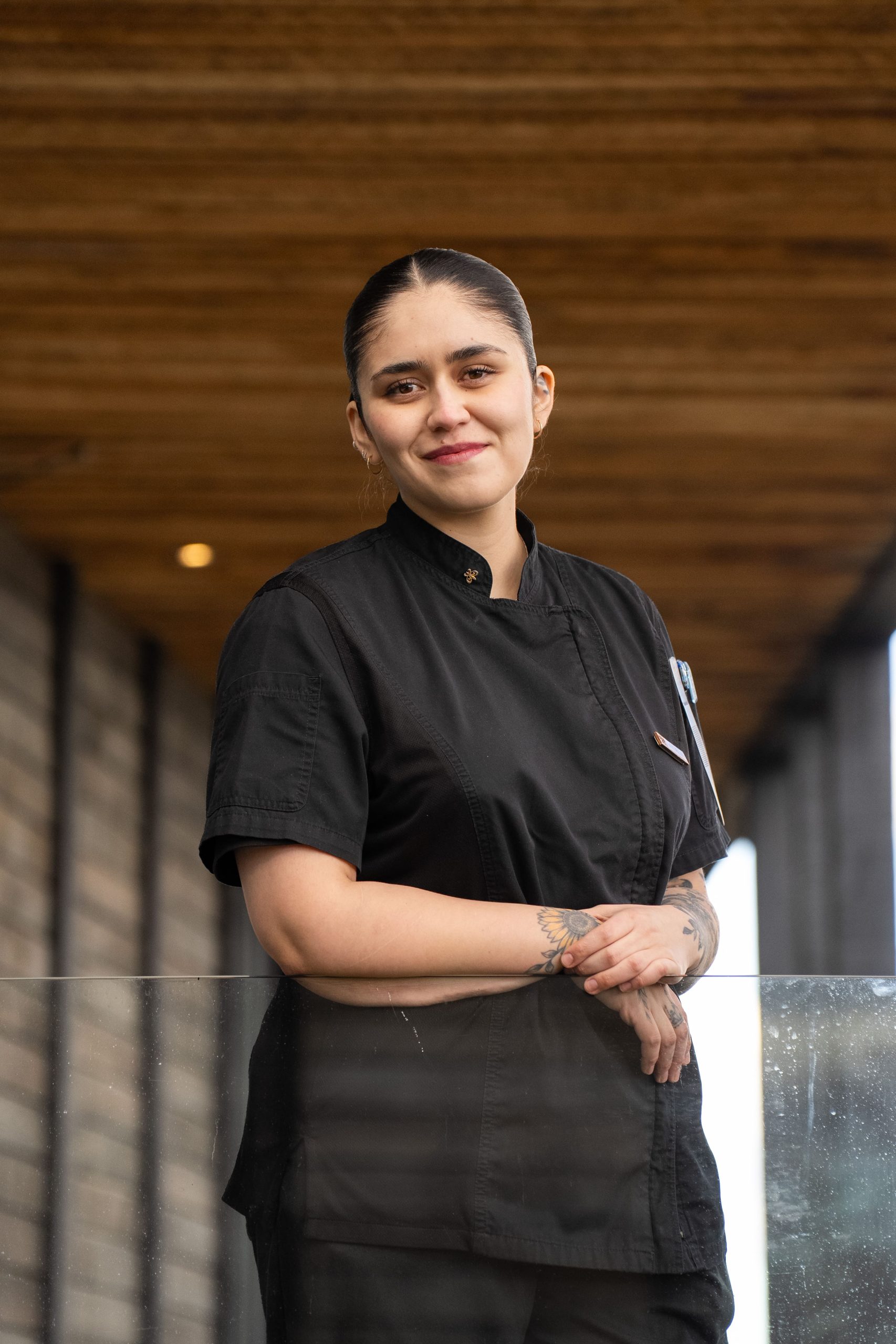 Portrait Of A Smiling Hospitality Staff Member In Black Uniform Standing Outdoors Beside A Glass Railing.