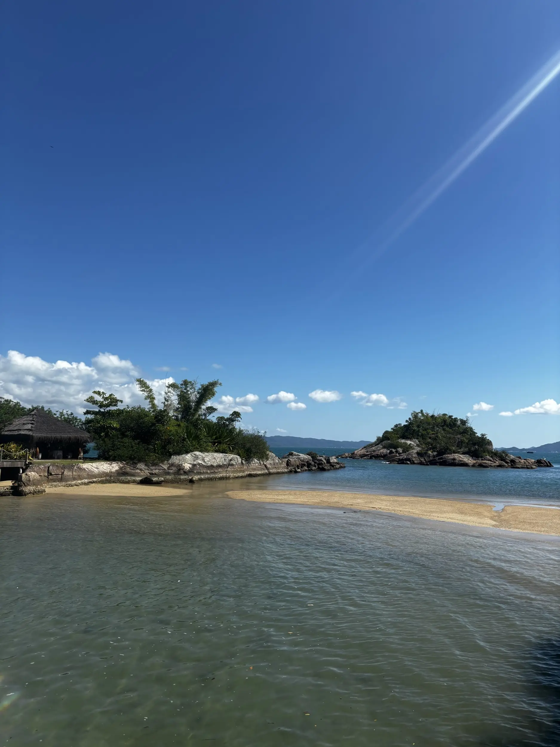 Island coastline with clear water, a sandy beach, and small rocky islets under a bright blue sky.